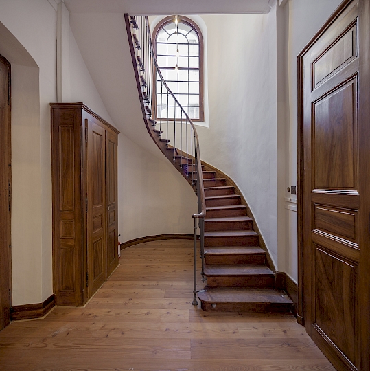 Vue générale avant travaux des escaliers balancés donnant accès aux galeries de l’étage supérieur (nord-est). Le parquet du début des années 2000, collé sur un panneau aggloméré, a été déposé et remplacé par un parquet en mélèze brossé à l’ancienne (type Admonters). De nouveaux luminaires en laiton ont été installés, un lustre Poulsen dans l’accès et une lampe sur mesure dans la montée des escaliers. Ponçage et huilage des marches en chêne. Ponçage et peinture MLE4 du barreaudage. Le tableau électrique secondaire a été installé dans l’armoire récupérée et restaurée de la galerie. Photo après travaux.