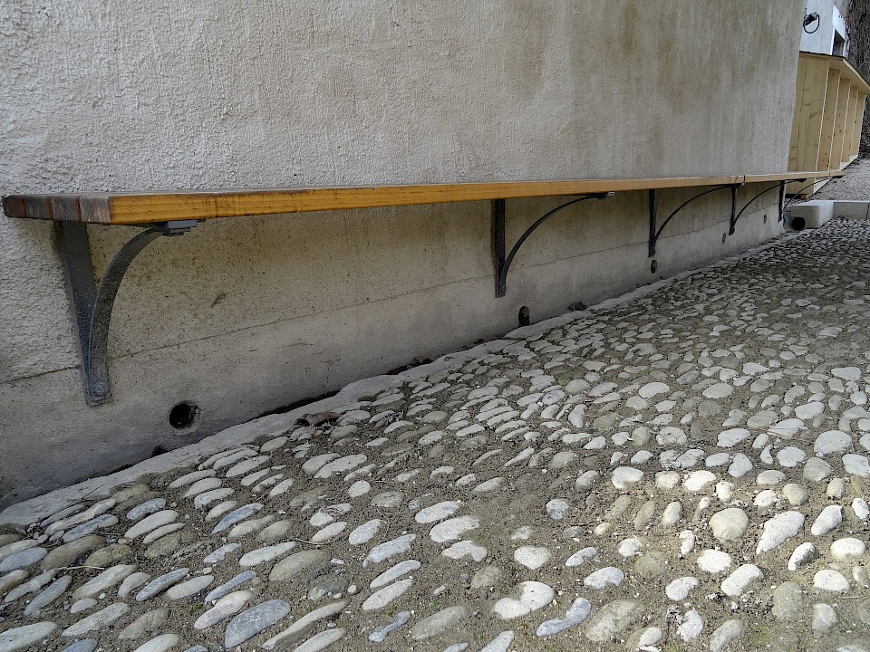 Mise en place d'une calade au même niveau que celle d'origine. Réfection du crépi du mur d'appui du couvert avec un mortier de chaux hydratée et chaux hydraulique. Confection d'une plinthe enrichie de ciment. Mise en place de bancs avec consoles en fer.