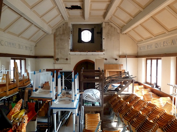 Vue d'ensemble de la nef en direction de l'entrée. Le canal électrique en bois situé au dessus des fenêtres sera déposé et intégré au futur plancher. Les deux versets bibliques ainsi que le caisson perforé en toile seront restaurés. En vue du changement de fonction de la nef, une mezzanine faisant office d'espace d'archivage est projetée dans le prolongement de la galerie en partie supérieur de la nef. L'accès s'y fera par le beffroi. La structure de la mezzanine sera conçue de manière à contribuer à la consolidation de la Chapelle, avec l’incorporation de tirants métalliques.
